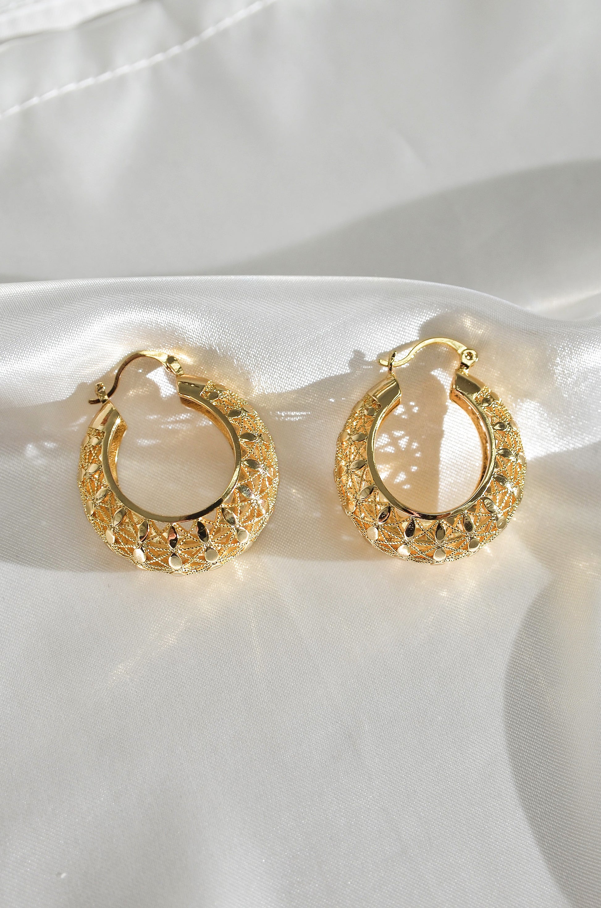 A pair of gold-colored filigree hoop earrings with a woven-inspired design, displayed against a white fabric background. by DBL Jewelry