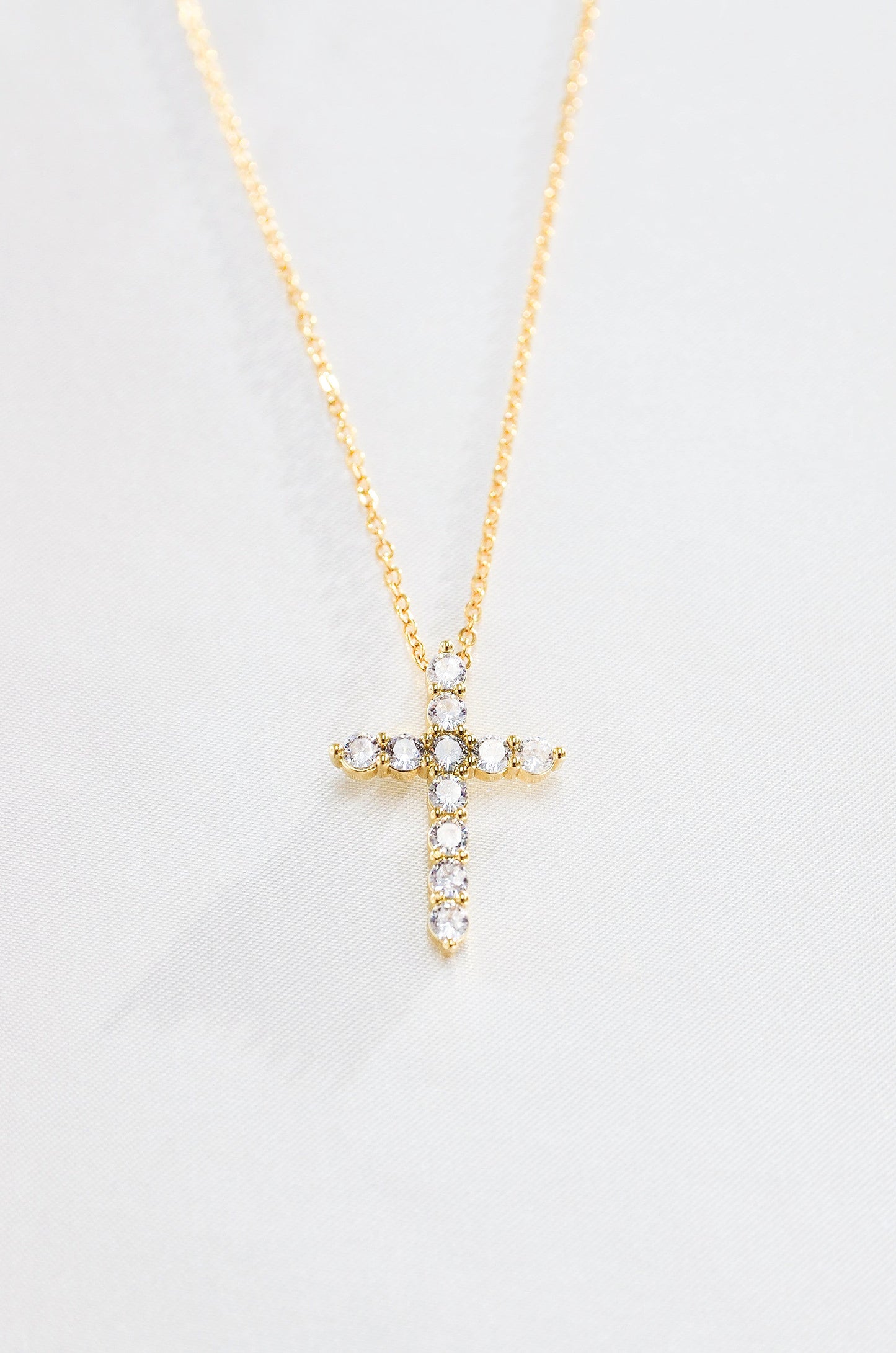 A gold-colored cross necklace with cubic zirconia stones, displayed against a white background. By DBL Jewelry