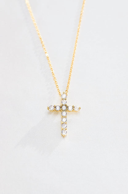 A gold-colored cross necklace with cubic zirconia stones, displayed against a white background. By DBL Jewelry