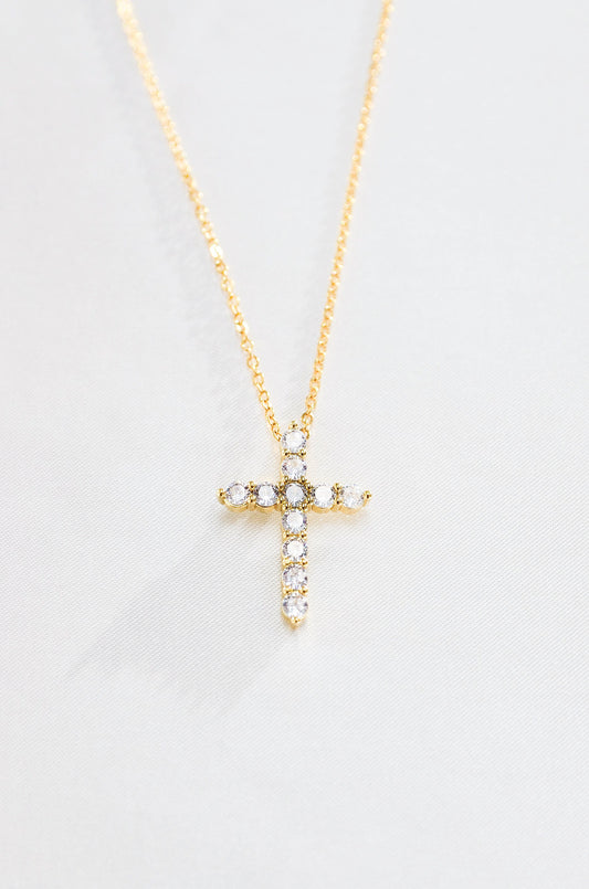 A gold-colored cross necklace with cubic zirconia stones, displayed against a white background. By DBL Jewelry