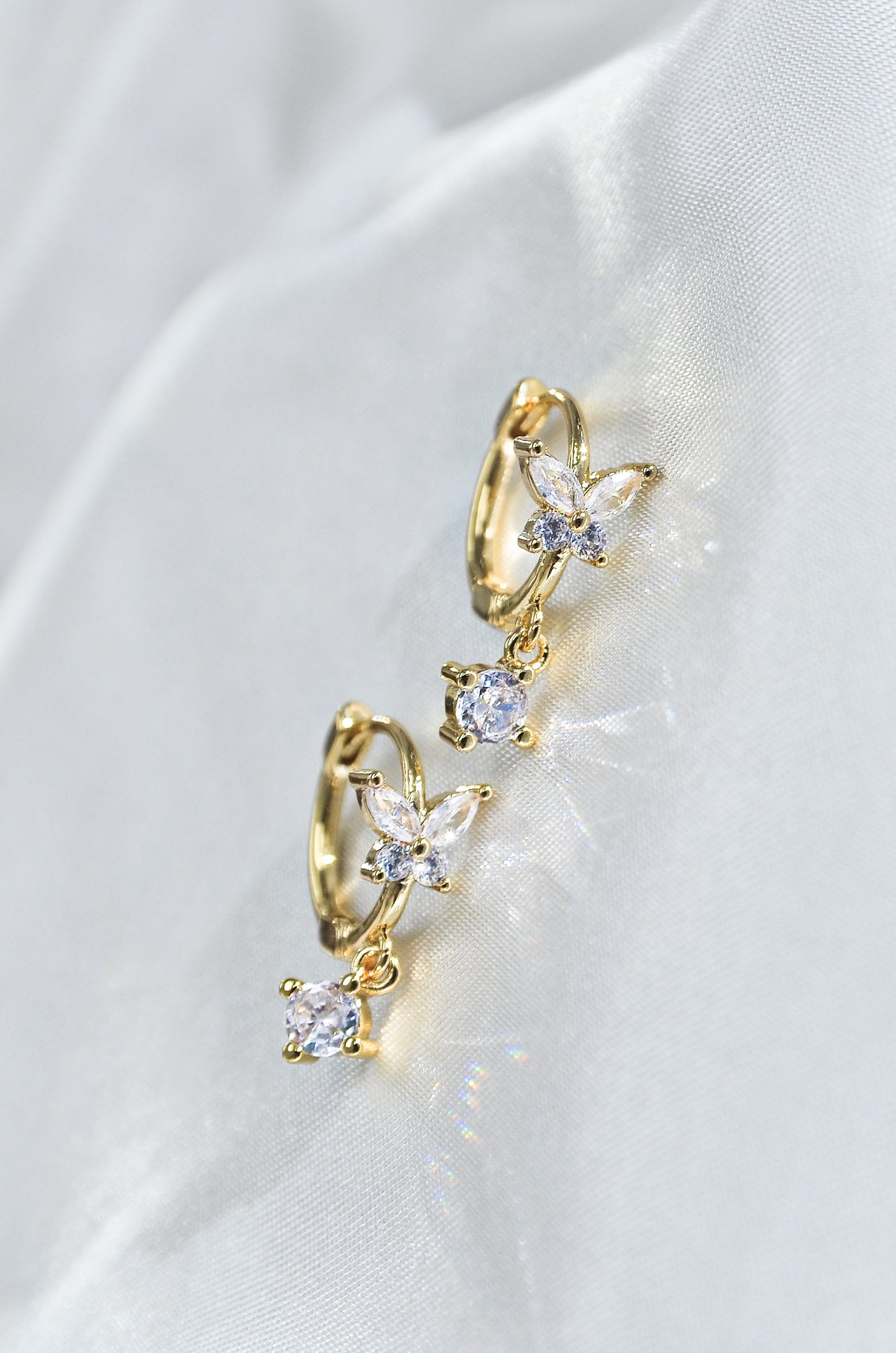 A pair of dainty butterfly-shaped drop earrings with gold filling and sparkling diamonds, displayed on a white fabric. By DBL Jewelry