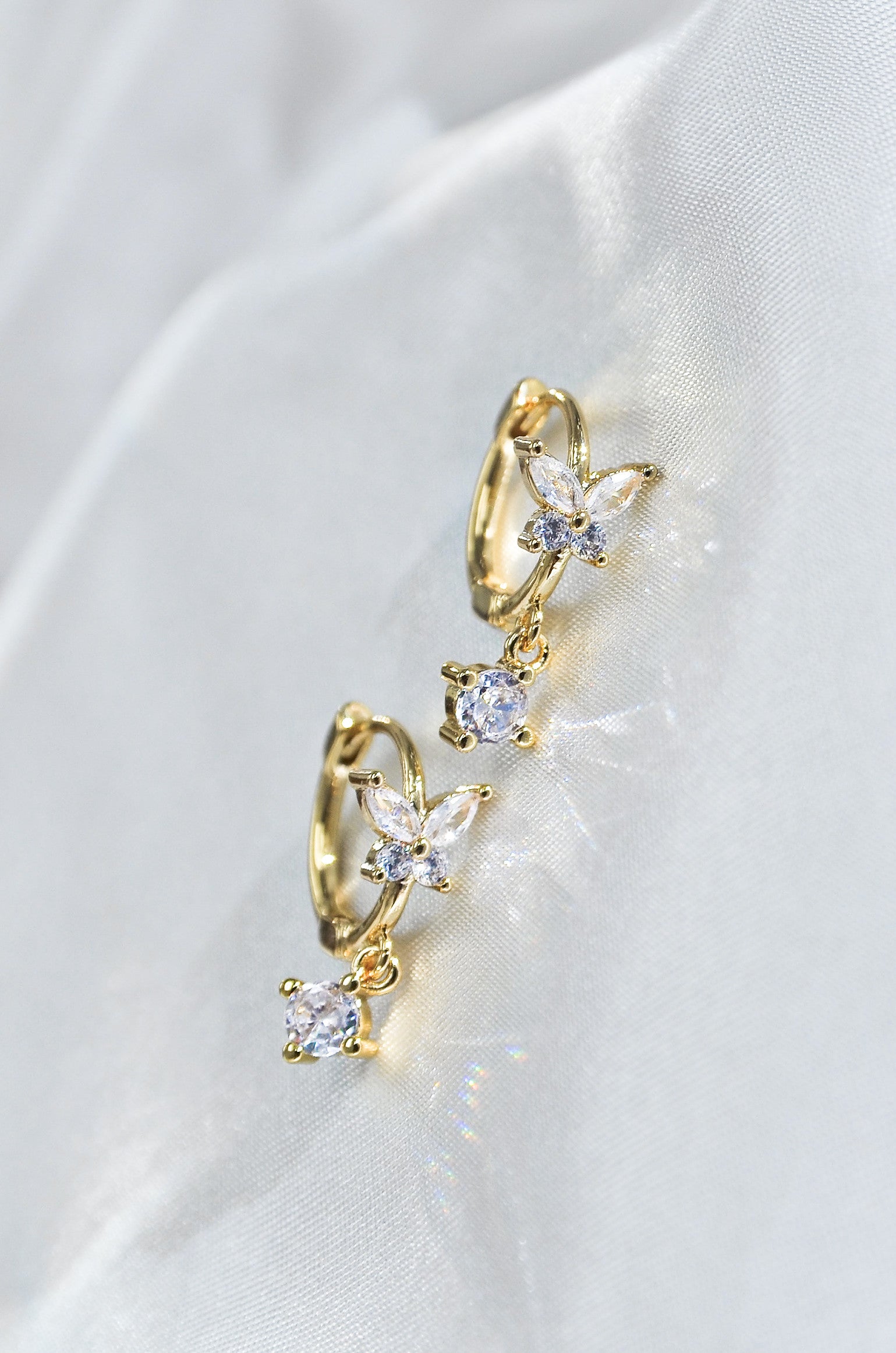 A pair of dainty butterfly-shaped drop earrings with gold filling and sparkling diamonds, displayed on a white fabric. By DBL Jewelry