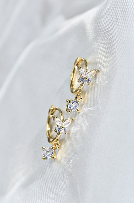 A pair of dainty butterfly-shaped drop earrings with gold filling and sparkling diamonds, displayed on a white fabric. By DBL Jewelry