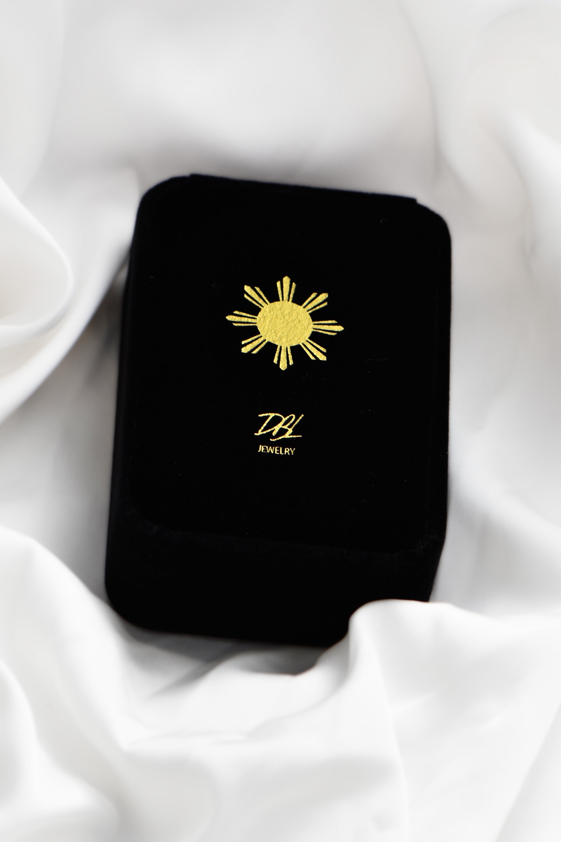 Black jewelry box with a yellow sun design and 'DBL' logo on a white fabric background