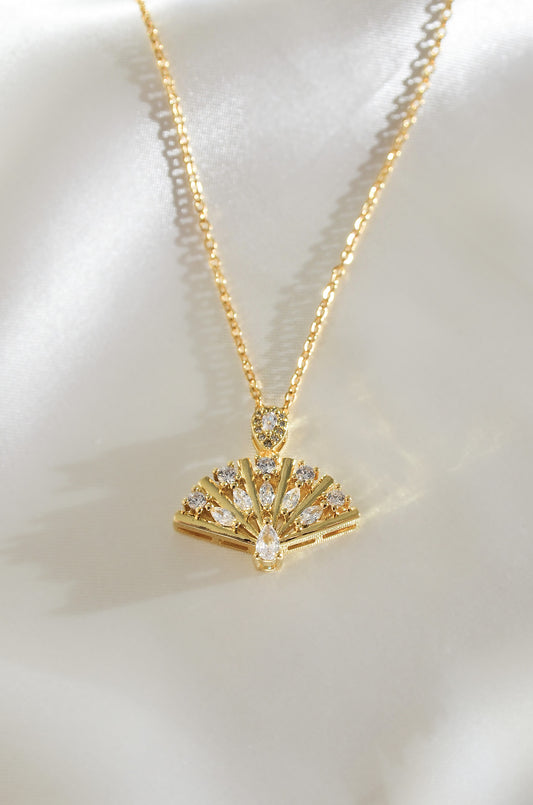A gold-colored necklace featuring a fan-shaped pendant adorned with sparkling cubic zirconia stones, displayed on a silky background. by DBL Jewelry