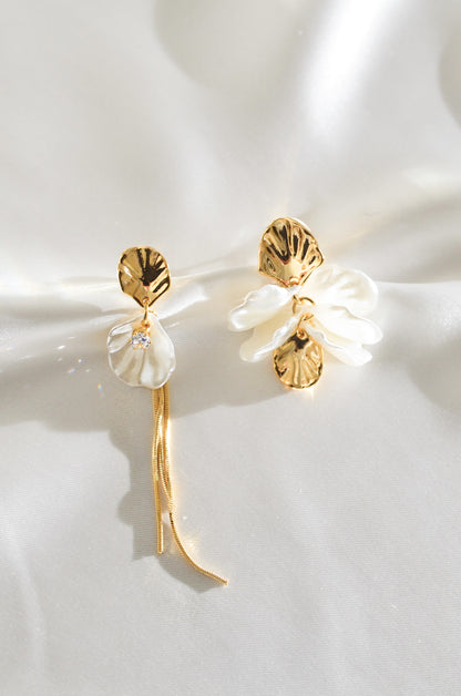 A pair of gold-colored earrings featuring white pearl drops and floral designs, displayed on a silky background. By DBL Jewelry