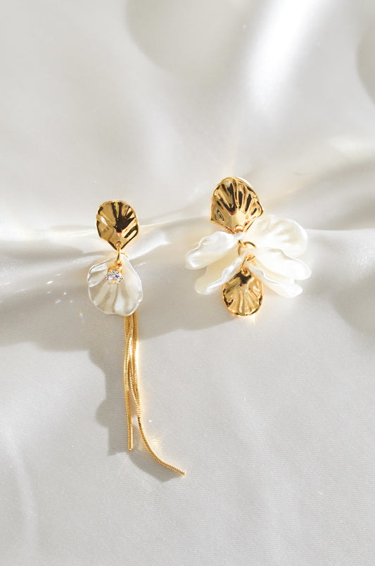 A pair of gold-colored earrings featuring white pearl drops and floral designs, displayed on a silky background. By DBL Jewelry